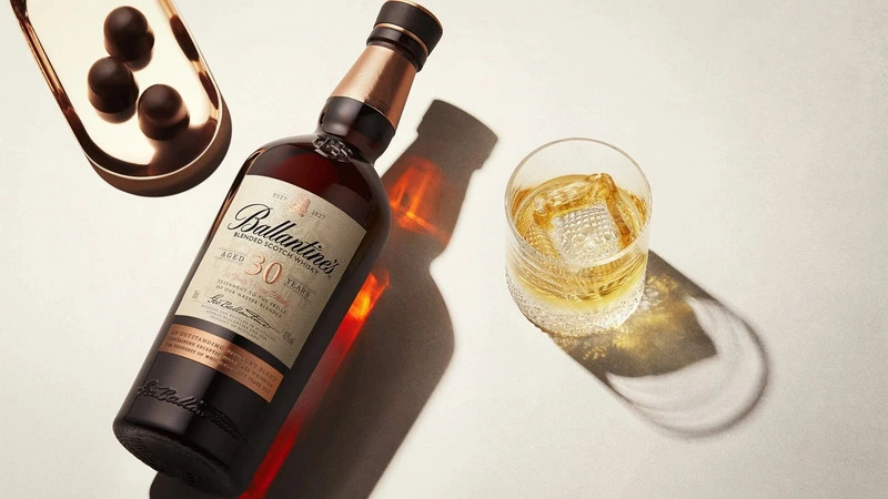 Chai rượu Ballantines nằm trên mặt phẳng và cốc rượu có đá bên cạnh